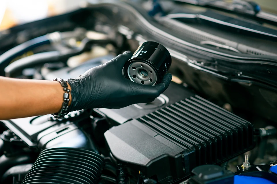 Main d'un mécanicien automobile portant des gants de protection noirs tenant un filtre à huile neuf au-dessus du compartiment moteur d'un véhicule lors d'une opération de maintenance