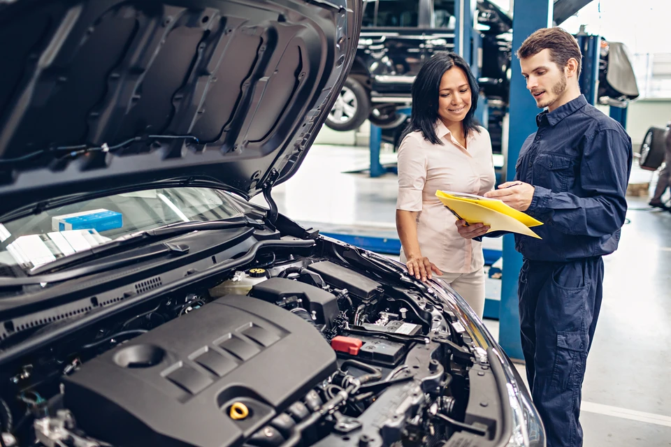 Atelier automobile moderne avec mécanicien en combinaison bleue consultant documents jaunes avec cliente en chemisier beige près d'un véhicule noir au capot ouvert